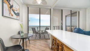 Una habitación moderna con una gran mesa blanca, sillas negras y un taburete de mimbre. Cuenta con un ventanal con vistas al océano.