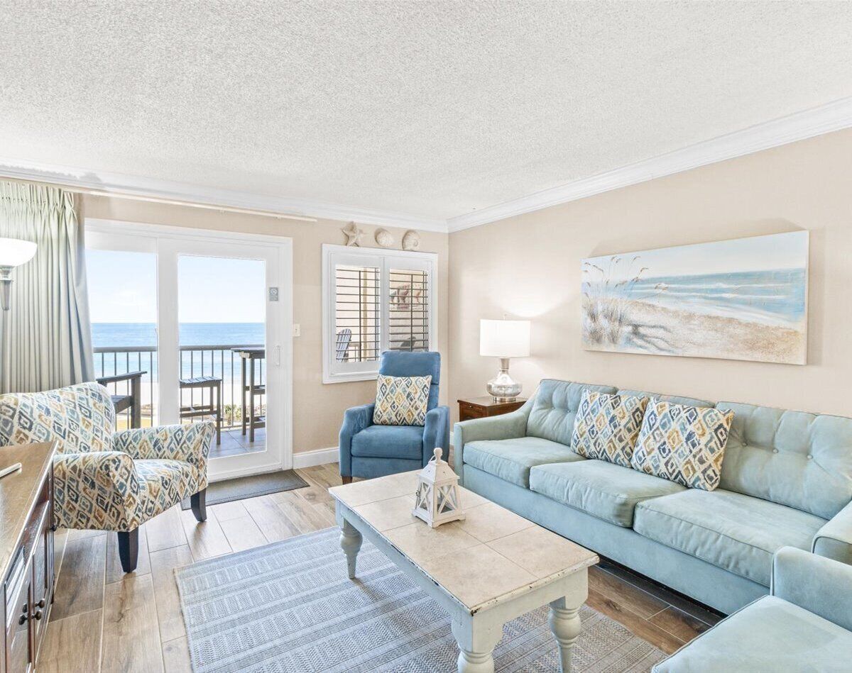 A living room with a view of the ocean, a blue armchair, a light blue sofa, and a wooden coffee table.
