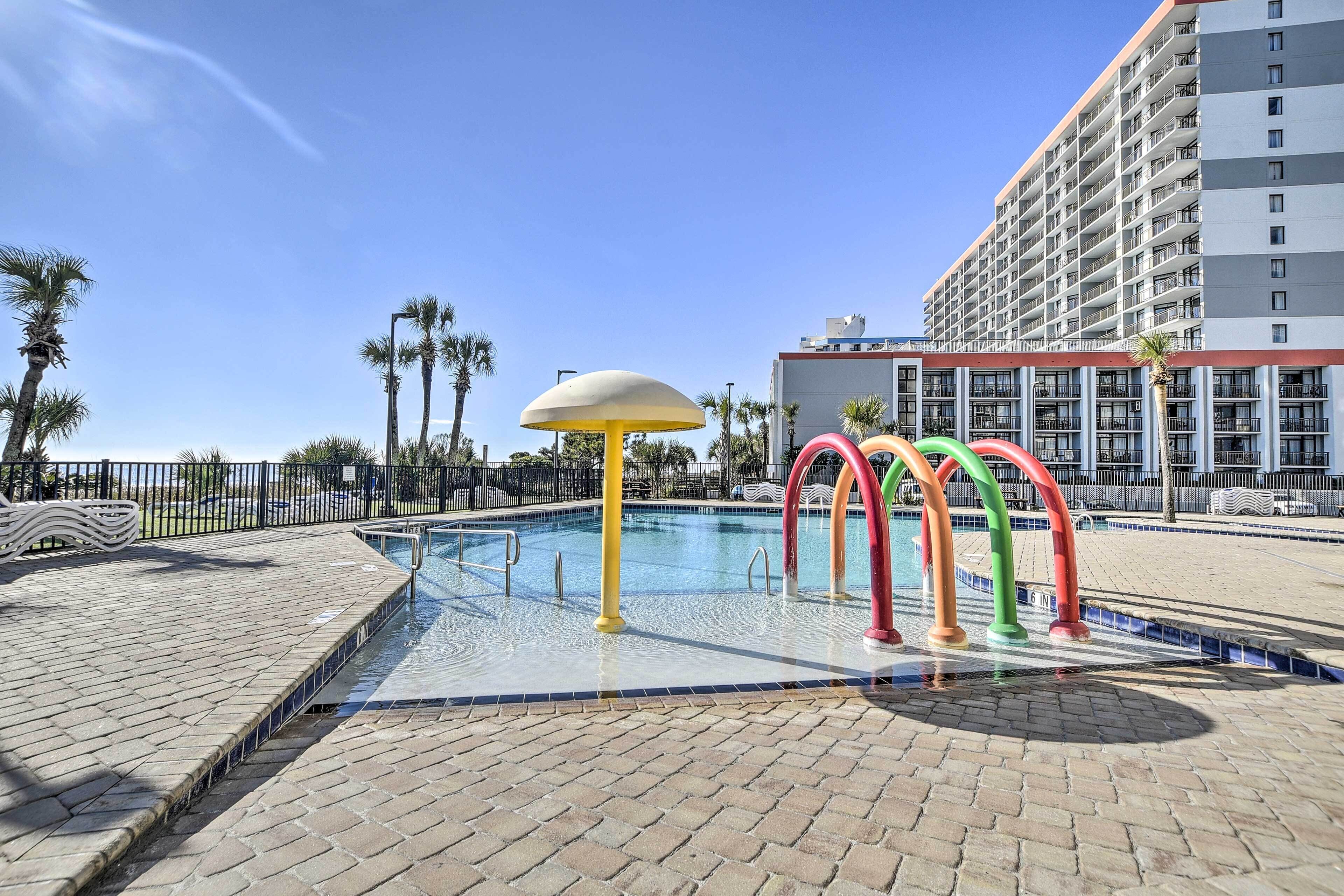 A swimming pool with colorful arches, a palm tree-lined walkway, and a hotel building in the background.