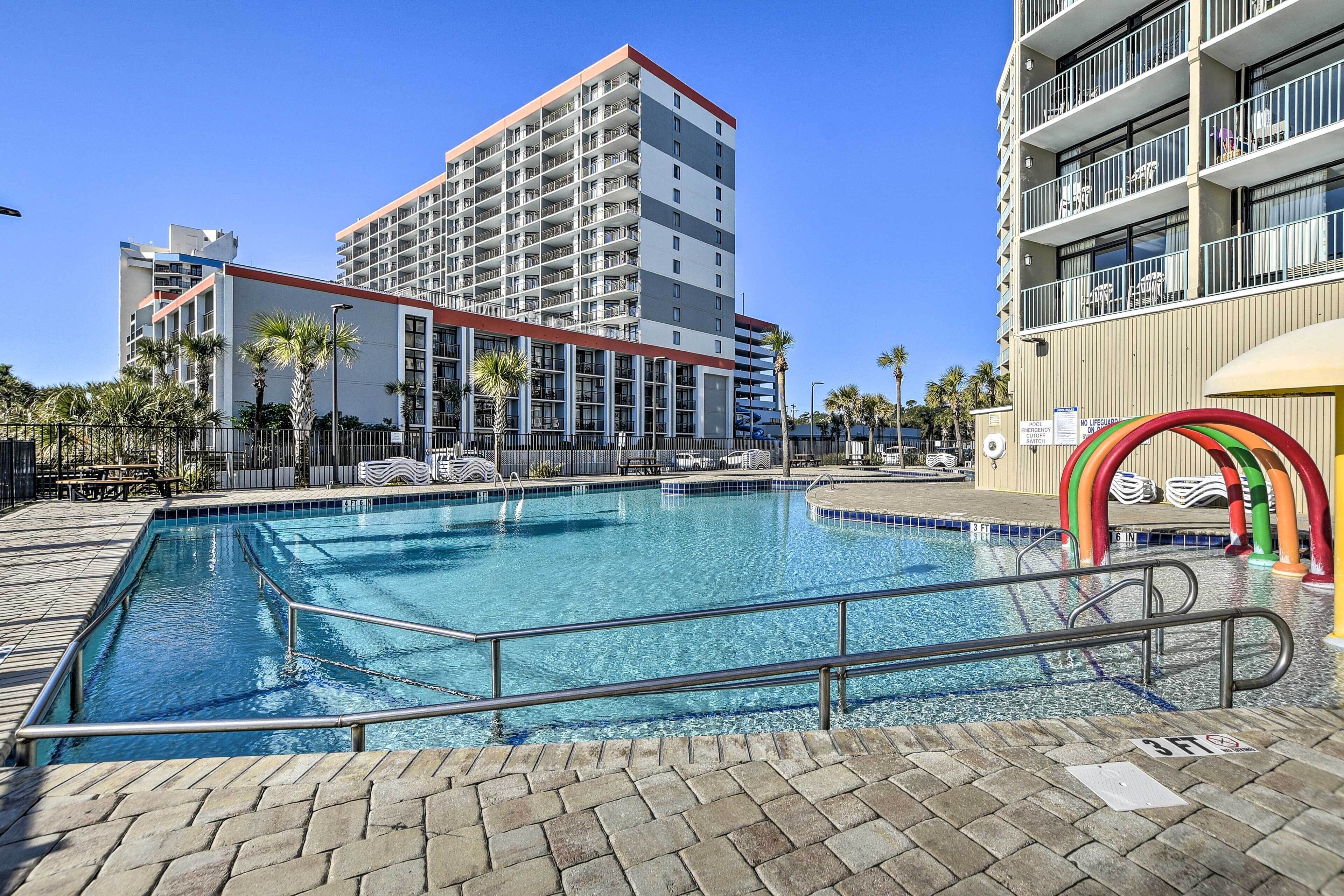 Piscina com deck ladrilhado, rodeada por palmeiras e um edifício com varandas.