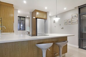 Una cocina moderna con encimera blanca, muebles de madera y un refrigerador grande. Hay dos taburetes de bar y una ventana con persianas.