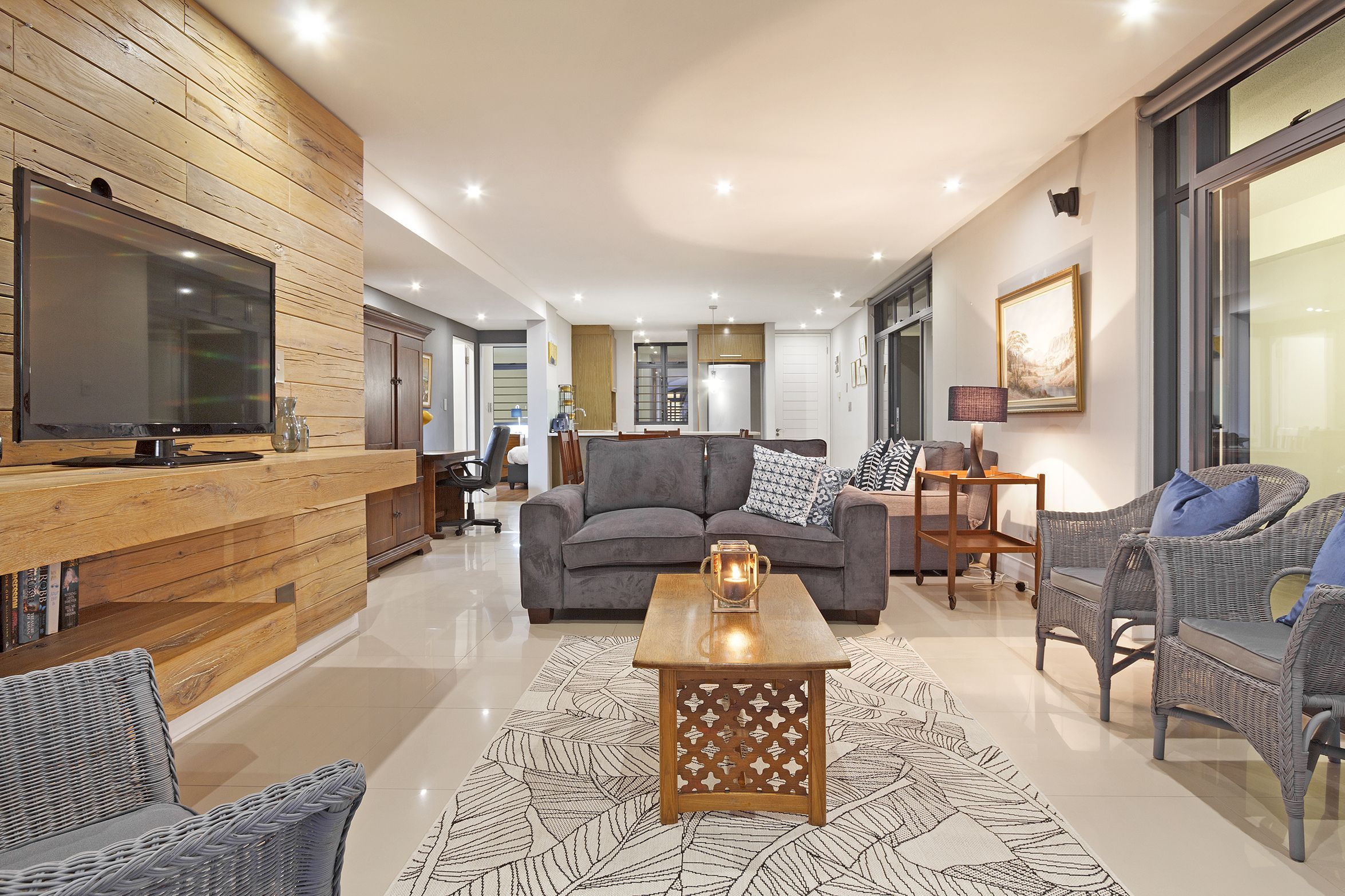 A modern living room with a wooden accent wall, a large flat-screen TV, a grey sofa, wicker chairs, and a patterned rug.