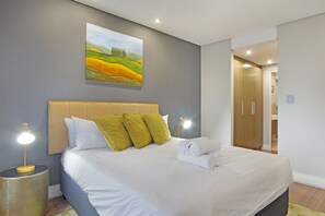 A neatly made bed with white linens and green pillows, a bedside lamp, and a painting of a landscape on the wall. - Eden on the Bay 171 (Cape Town)