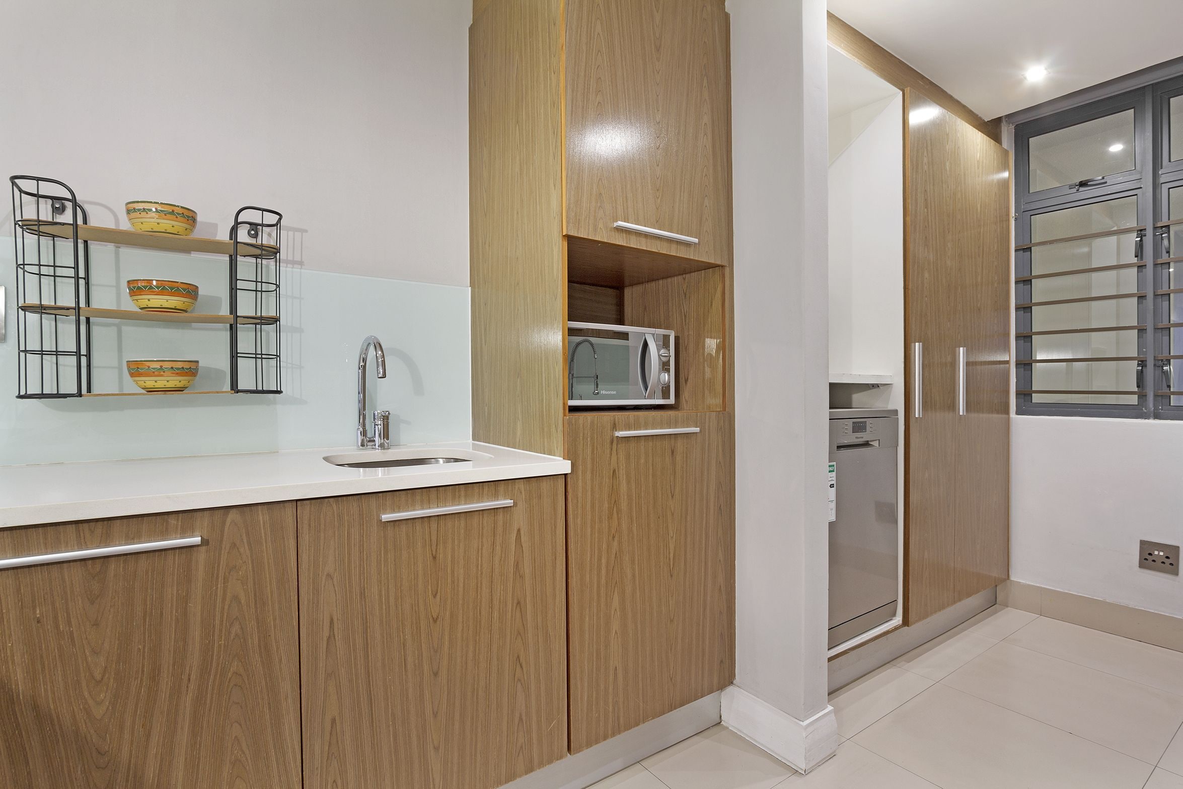 A modern kitchen with wooden cabinets, a white countertop, a built-in microwave, and a glass-fronted dishwasher.