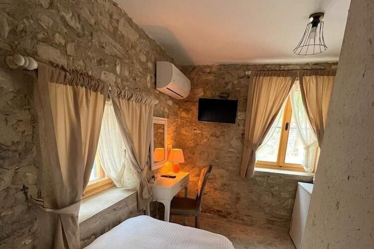 A bedroom with stone walls, a bed, a desk with a chair, a TV, and a window with curtains.
