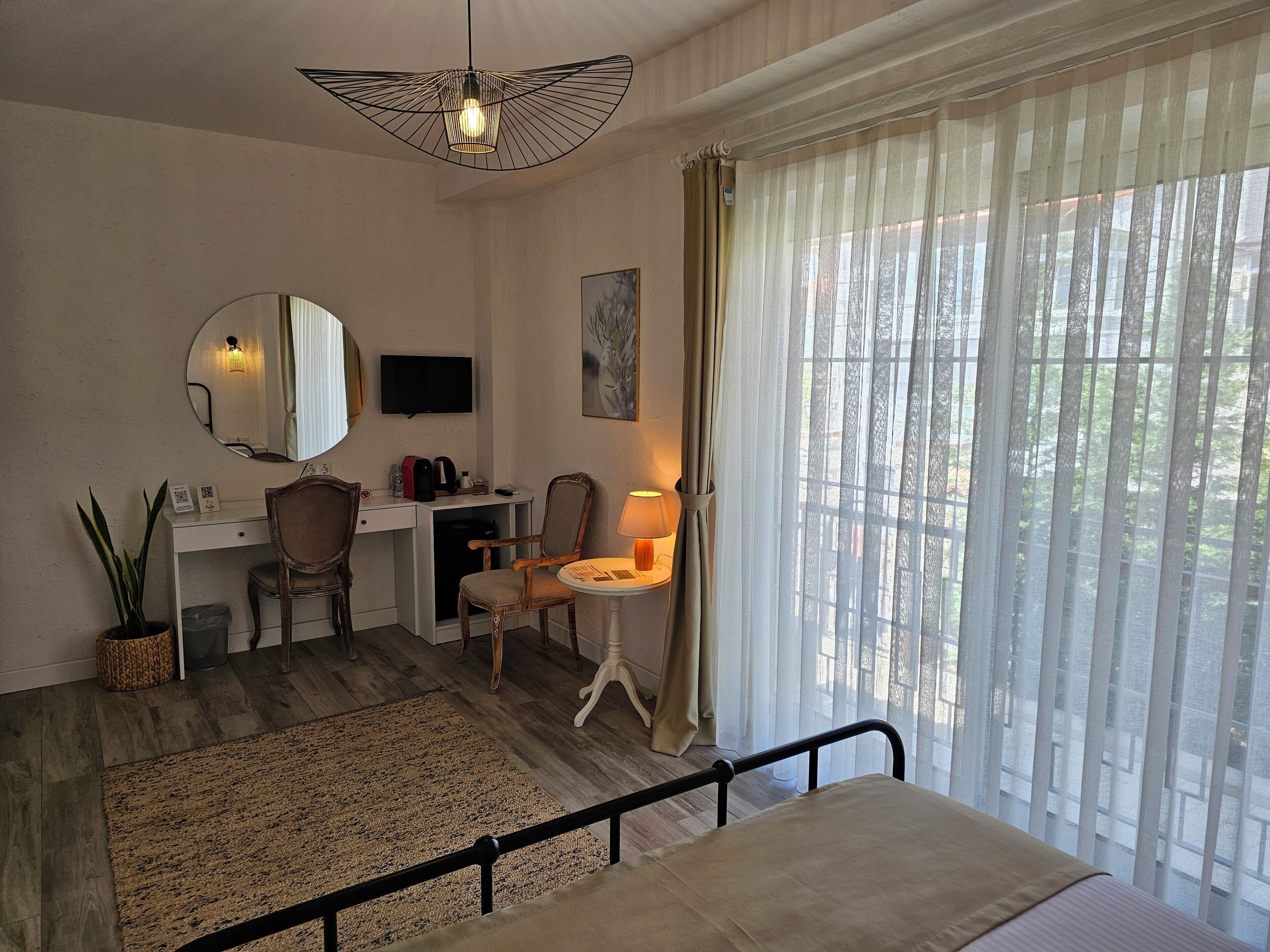 A hotel room with a bed, a desk with a chair, a TV, a lamp, and a large window with curtains.