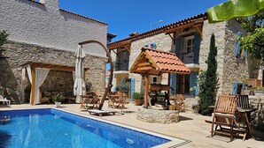 Una piscina con tumbonas y una pérgola, rodeada de edificios de piedra con contraventanas azules.