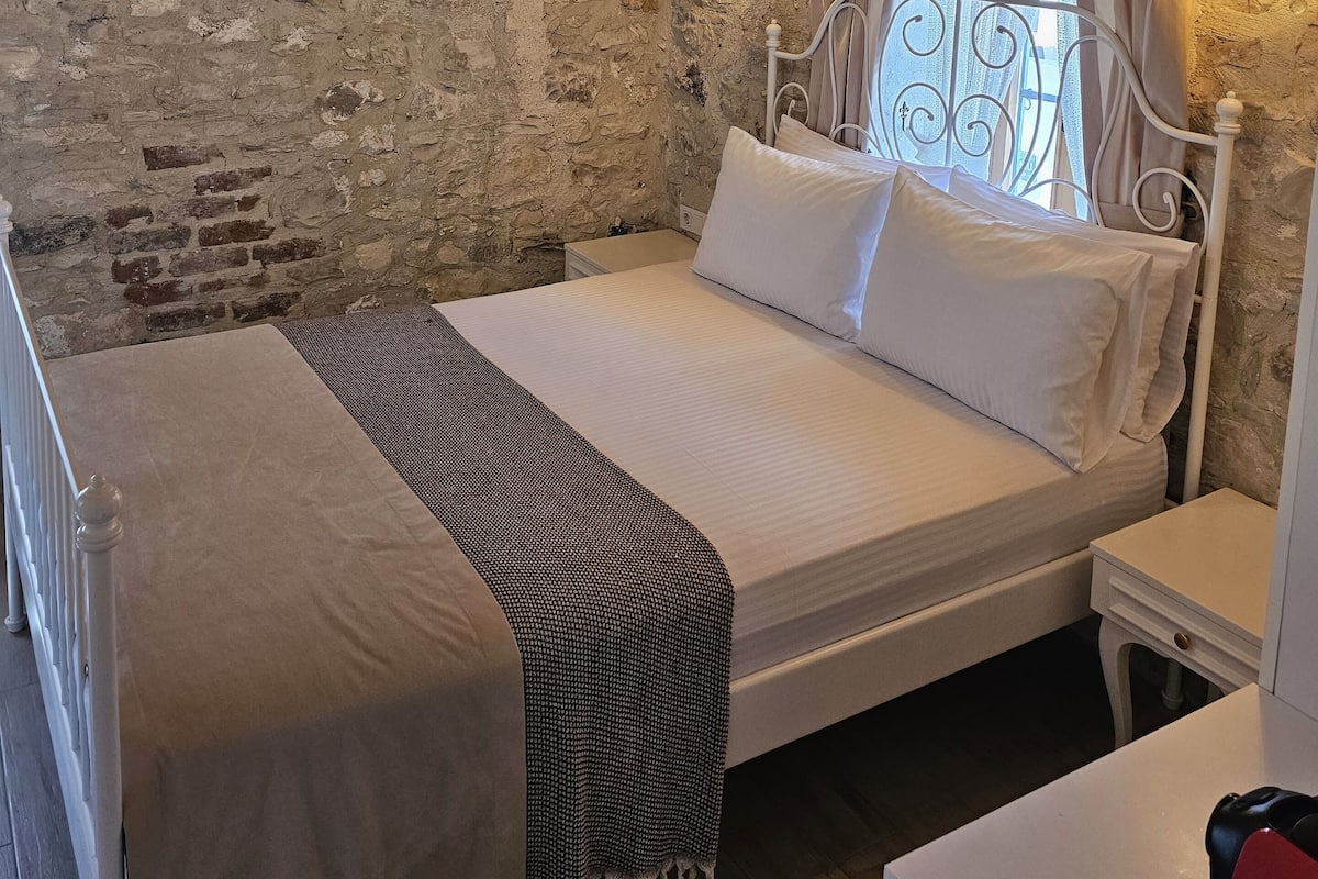 A bedroom with a stone wall, a bed with white linens, a chandelier, and a window with curtains.
