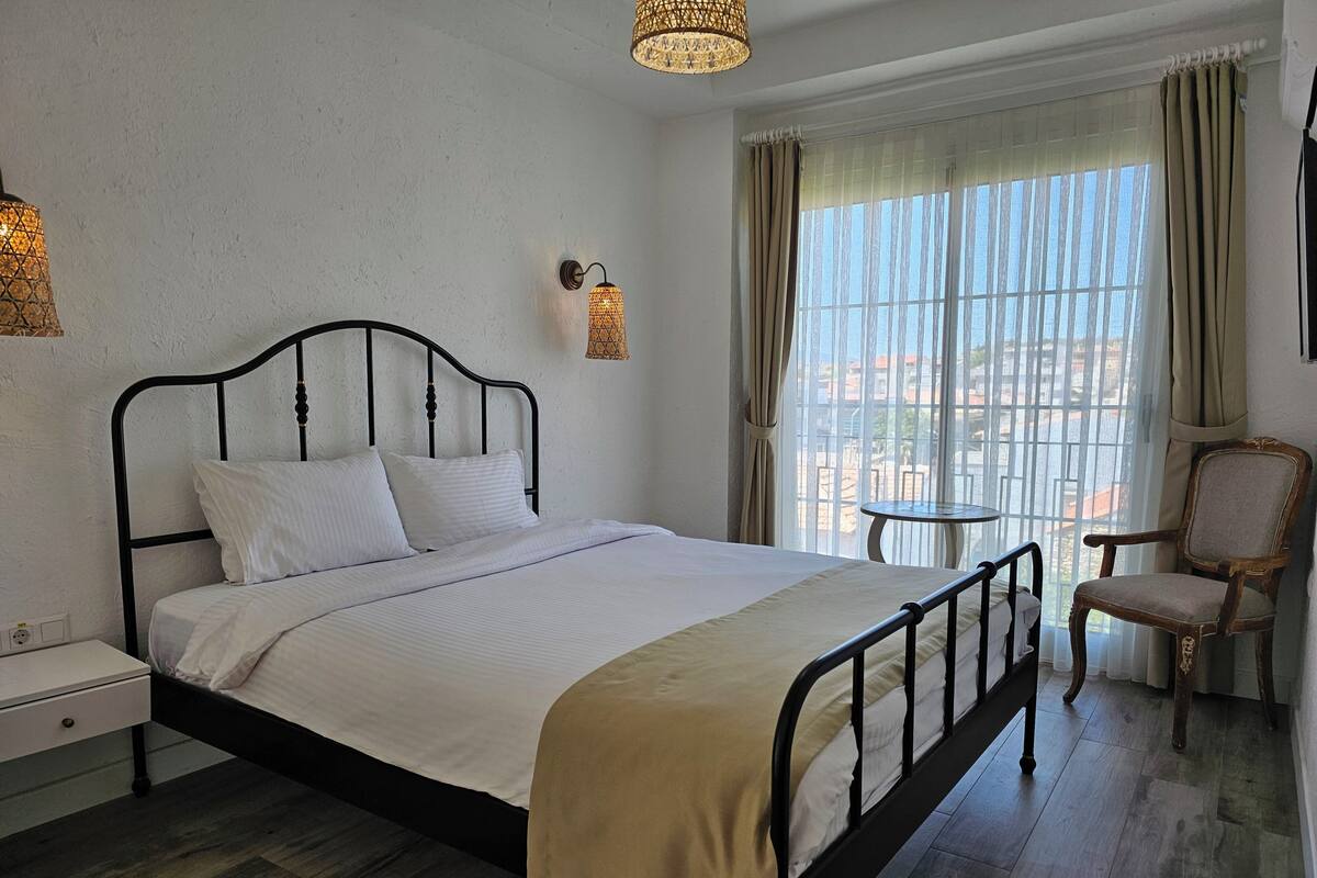 A neatly made bed with a headboard, a bedside table, a chair, and a window with curtains.