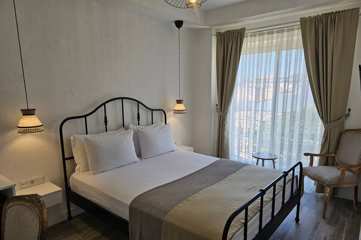 A neatly arranged bedroom with a bed, bedside lamps, a chair, and a view of the outdoors through a window.