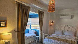 Un dormitorio con cama, mesita de noche con lámpara, ventana con cortinas, lámpara colgante y unidad de aire acondicionado.