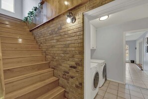 A staircase with wooden steps leading to a brick wall, a washing machine, and a white sink in a laundry area. - Luxury Home 2-Mins to Downtown w/Sauna+Plunge Pool (Fredericksburg)