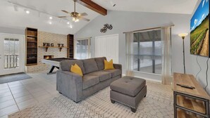 A modern living room with a grey sofa, ottoman, and a pool table. There is a ceiling fan, a fireplace, and a large window with blinds. - Luxury Home 2-Mins to Downtown w/Sauna+Plunge Pool (Fredericksburg)