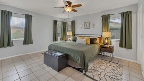 A bedroom with a bed, a ceiling fan, a nightstand with a lamp, a bench, and a window with curtains. - Luxury Home 2-Mins to Downtown w/Sauna+Plunge Pool (Fredericksburg)