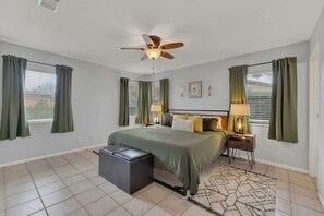 Une chambre à coucher avec un lit, un ventilateur de plafond, une table de chevet avec une lampe, un banc et une fenêtre avec des rideaux.
