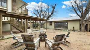 A covered outdoor seating area with wooden chairs and a fire pit, adjacent to a building with a balcony. - Luxury Home 2-Mins to Downtown w/Sauna+Plunge Pool (Fredericksburg)