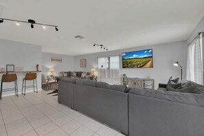 Un salon moderne avec un canapé d’angle gris, un coin repas avec deux tabourets de bar, une grande fenêtre avec des rideaux blancs et un tableau représentant un paysage accroché au mur.
