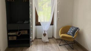 A room with a black wardrobe, a yellow chair, a white curtain, and a window with a view of greenery.