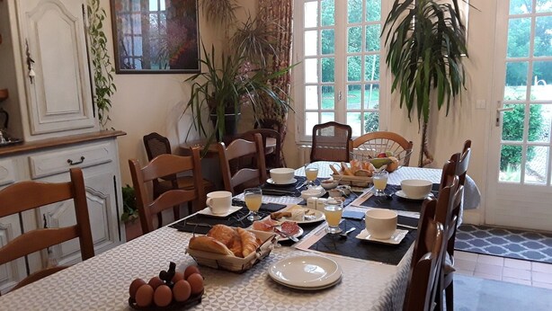 A dining area with a table set for breakfast, including bread, eggs, and fruit. There are chairs around the table, and a window with a view of greenery outside. - Chambres d'hôtes de l'île du saule (Saumur)