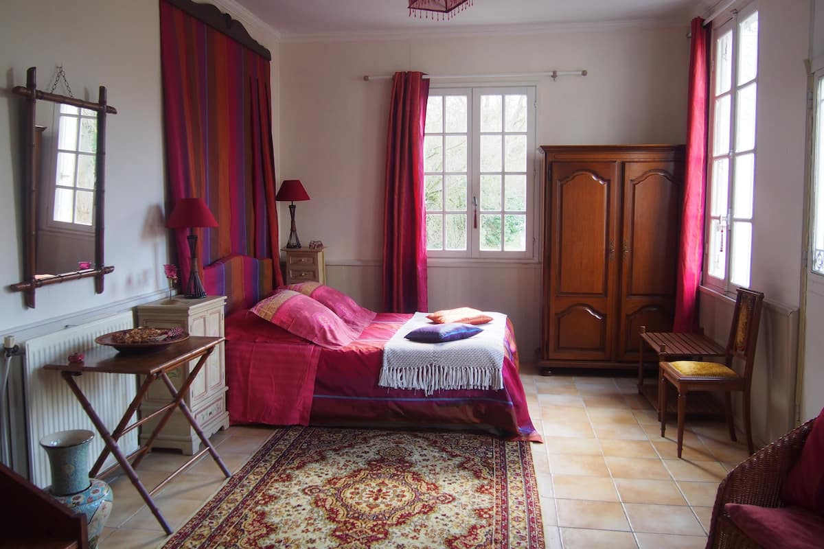 A bedroom with a bed, a wooden wardrobe, a chair, a table, and a window with curtains.