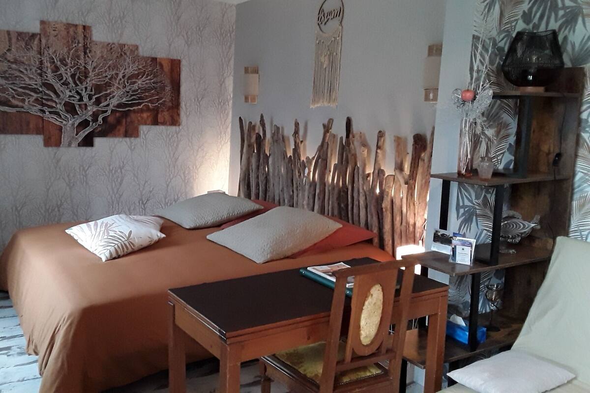 A bedroom with a bed, a wooden desk, and a chair. There is a wall with a tree artwork and a shelf with decorative items.