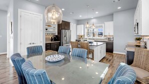 A modern kitchen with a central island, white cabinetry, and a dining area with striped chairs and a glass table.