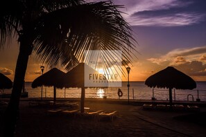 A beach scene with a sign for Fivesmar Property, palm trees, beach umbrellas, and lounge chairs.