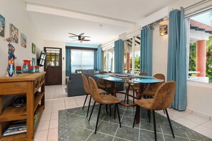 A dining area with a table and chairs, a ceiling fan, and a view of the ocean through large windows. - Diver's Paradise!  3 Bdrm True Oceanfront! 2A (San Miguel de Cozumel)