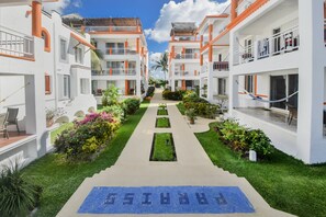 A hotel with a central pathway leading to a pool area, surrounded by lush greenery and white buildings with orange accents.