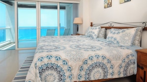 Quarto com uma cama grande, mesa de cabeceira, abajur e vista para o mar através de uma porta de correr.