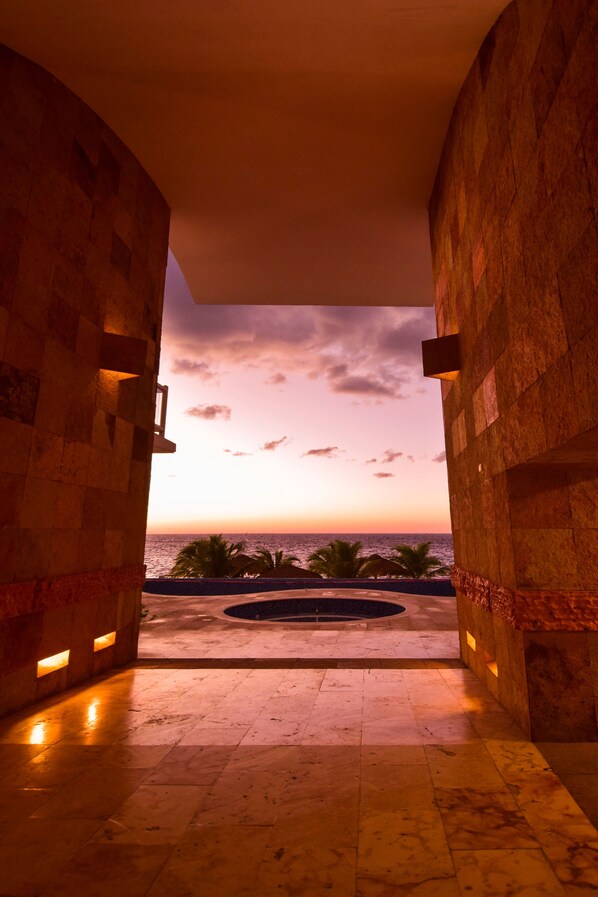 A view through a stone archway to a sunset over the ocean, with palm trees and a circular water feature in the foreground.
