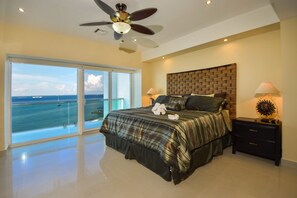 A bedroom with a large bed, a ceiling fan, a nightstand, and a view of the ocean.