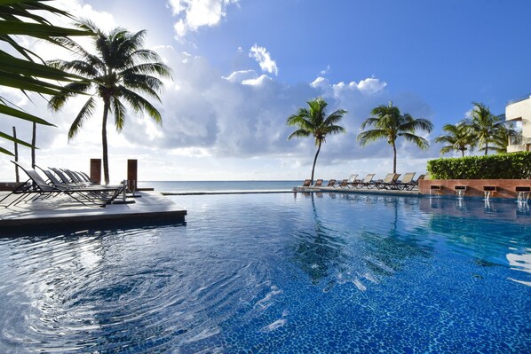 Une piscine à l’eau d’un bleu cristallin, entourée de palmiers et de chaises longues, avec vue sur une plage.