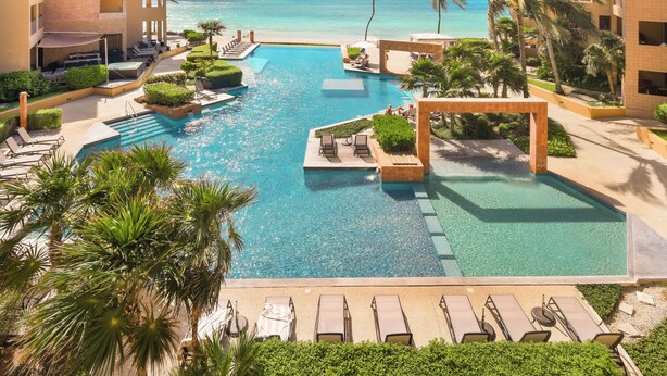 Ein Poolbereich in einem Resort mit klarem, blauem Wasser, Palmen und Blick auf den Ozean.