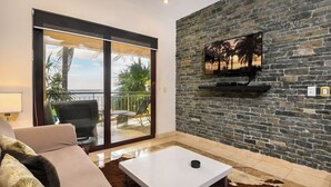 Un soggiorno con un divano, un tavolino, una TV montata su una parete in pietra e una porta scorrevole in vetro che dà su un balcone con palme.