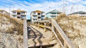 Una pasarela de madera que lleva a edificios frente al mar con balcones, rodeada de arena y vegetación escasa.