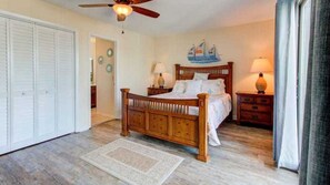 A bedroom with a wooden bed, two bedside tables with lamps, a ceiling fan, and a view of the sea through the window.