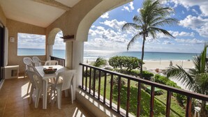 A balcony with a table and chairs, a view of the beach, and a palm tree.