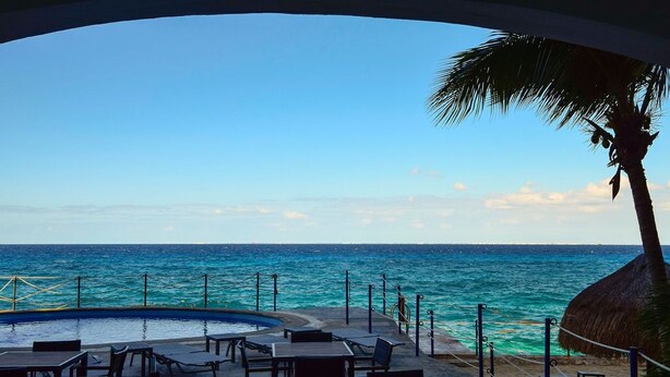 A poolside area with tables and chairs, a thatched-roof structure, and a view of the ocean. - Ground Floor 2 Bdrm Ocean Views! El Cantil LBN (San Miguel de Cozumel)