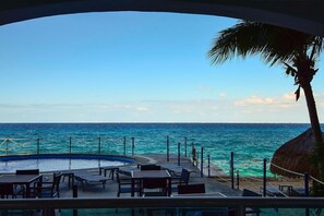 A poolside area with tables and chairs, a thatched-roof structure, and a view of the ocean.