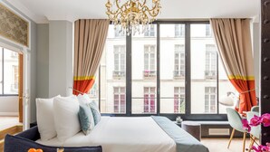 Ein Hotelzimmer mit einem großen Bett, einem fensterbelaubten Fenster, einer Kristallleuchtre und Blick auf Gebäude.