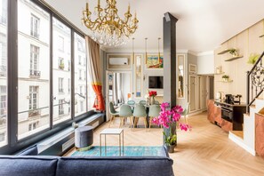 A modern living room with a large window, a chandelier, a dining area with chairs, and a kitchen with a stove. - Merveil - Luxury Suite - Louvre - Reaumur I (Paris)