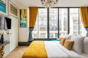 Habitación de hotel con una cama grande, televisión, mesita de mármol y vistas a edificios a través de amplias ventanas.