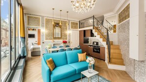 A spacious living room with a blue sofa, a chandelier, and a kitchen area with a staircase.
