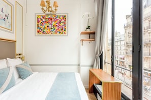 Habitación de hotel con cama, un cuadro abstracto de colores, una pequeña mesa de madera y vista a la ciudad a través de un ventanal grande.
