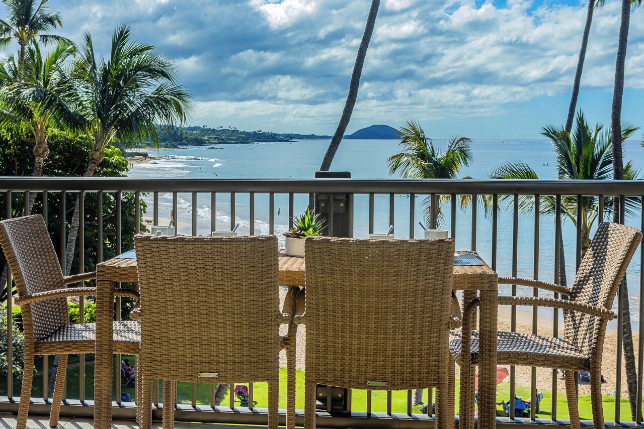 Un balcon avec des fauteuils en osier et une table, donnant sur une plage bordée de palmiers et offrant une vue sur l’océan.