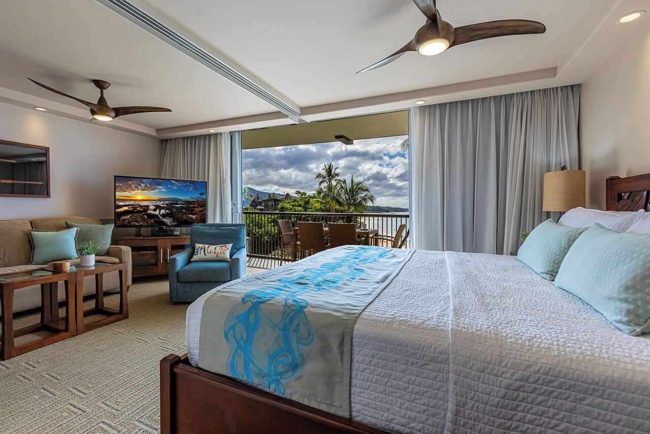 A hotel room with a large bed, a desk, a chair, a TV, and a balcony with a view of palm trees and a sunset.