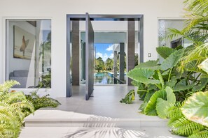 A modern house with a glass door leading to a pool, surrounded by lush greenery.