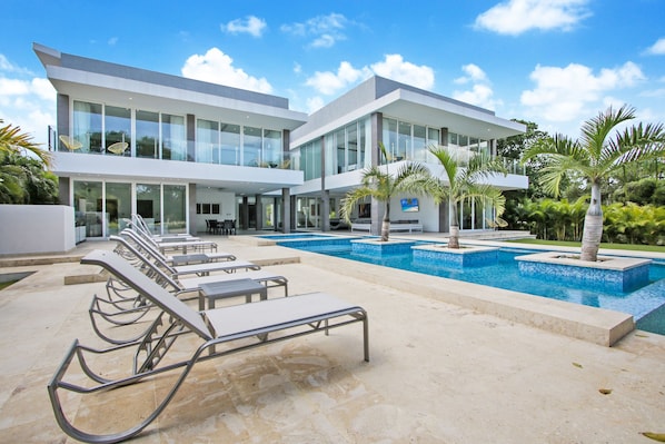 A modern two-story building with a swimming pool, lounge chairs, and palm trees.