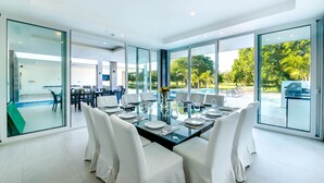 A modern dining area with a glass table, white chairs, and a view of a pool and greenery. - Ultra Modern Villa with Chef & Maid (Punta Cana)